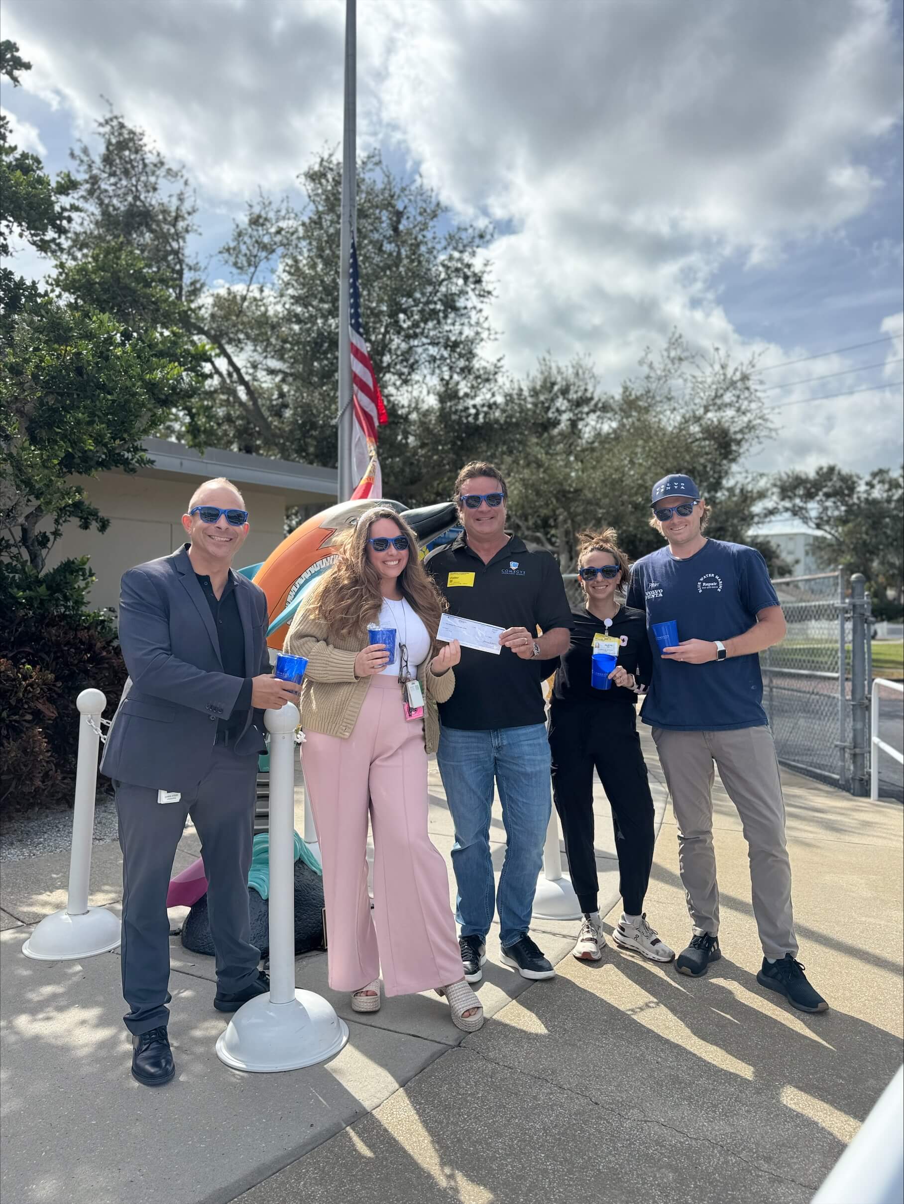 Five adults stand outside near a flagpole, holding blue ribbons and a check from Comegys Insurance Agency, with trees and a partly cloudy sky in the background.