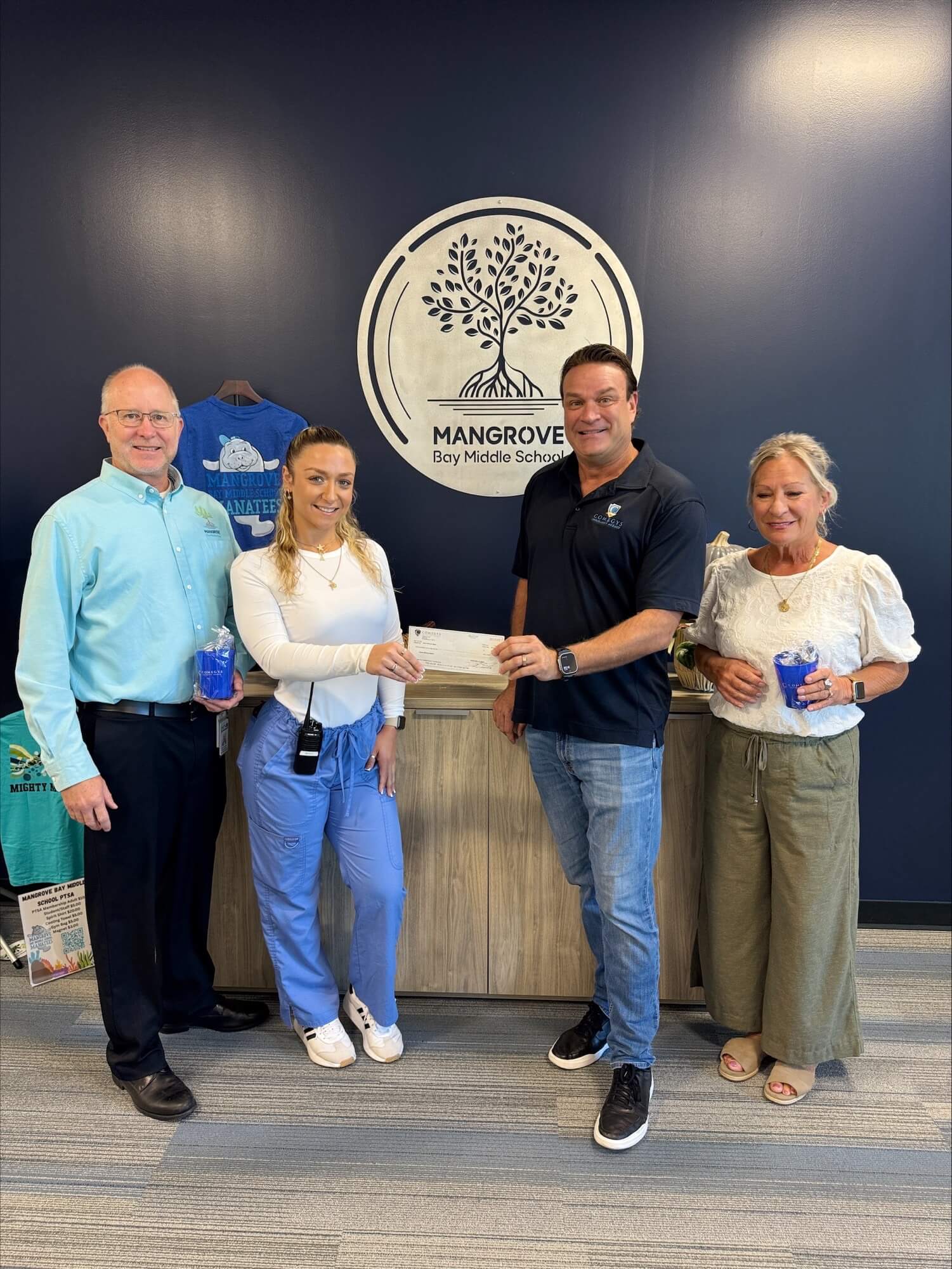 Four adults stand indoors in front of a Mangrove Bay Middle School sign; two are holding cups, and one is handing a check—sponsored by Comegys Insurance Agency—to another.