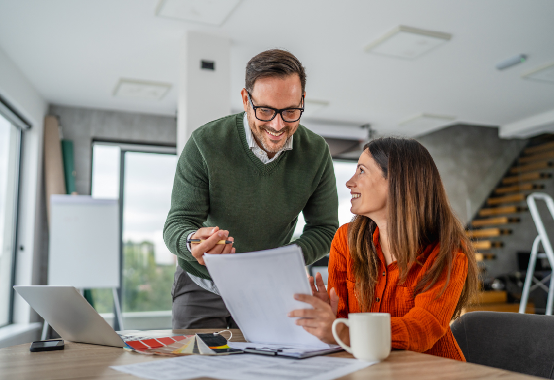 A man and a woman review documents together at a desk in a bright office, showing that thoughtful planning is their new love language, with a laptop, coffee mug, and papers spread out before them.
