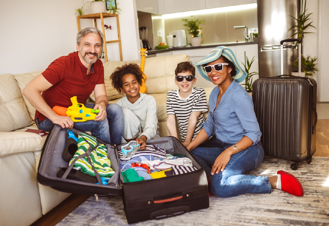 A family of four sits in a living room packing a suitcase with clothes and toys, using a travel checklist as they prepare for Spring Break travel.