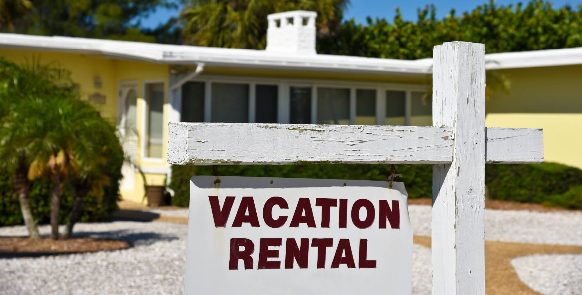 A white wooden sign reading "Vacation Rental" stands in front of a single-story yellow house with palm trees and gravel landscaping, highlighting the need for Florida Short Term Rental Insurance.