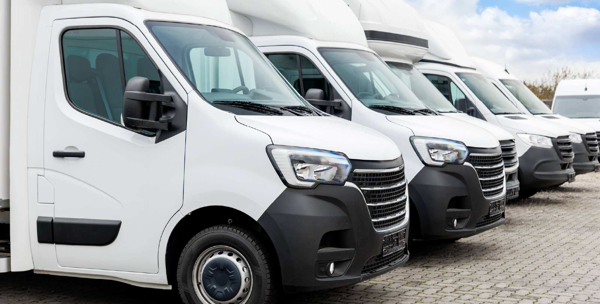 A row of white commercial delivery vans parked on a paved lot, ideal for contractors seeking reliable Commercial Automobile Insurance, with a partly cloudy sky in the background.