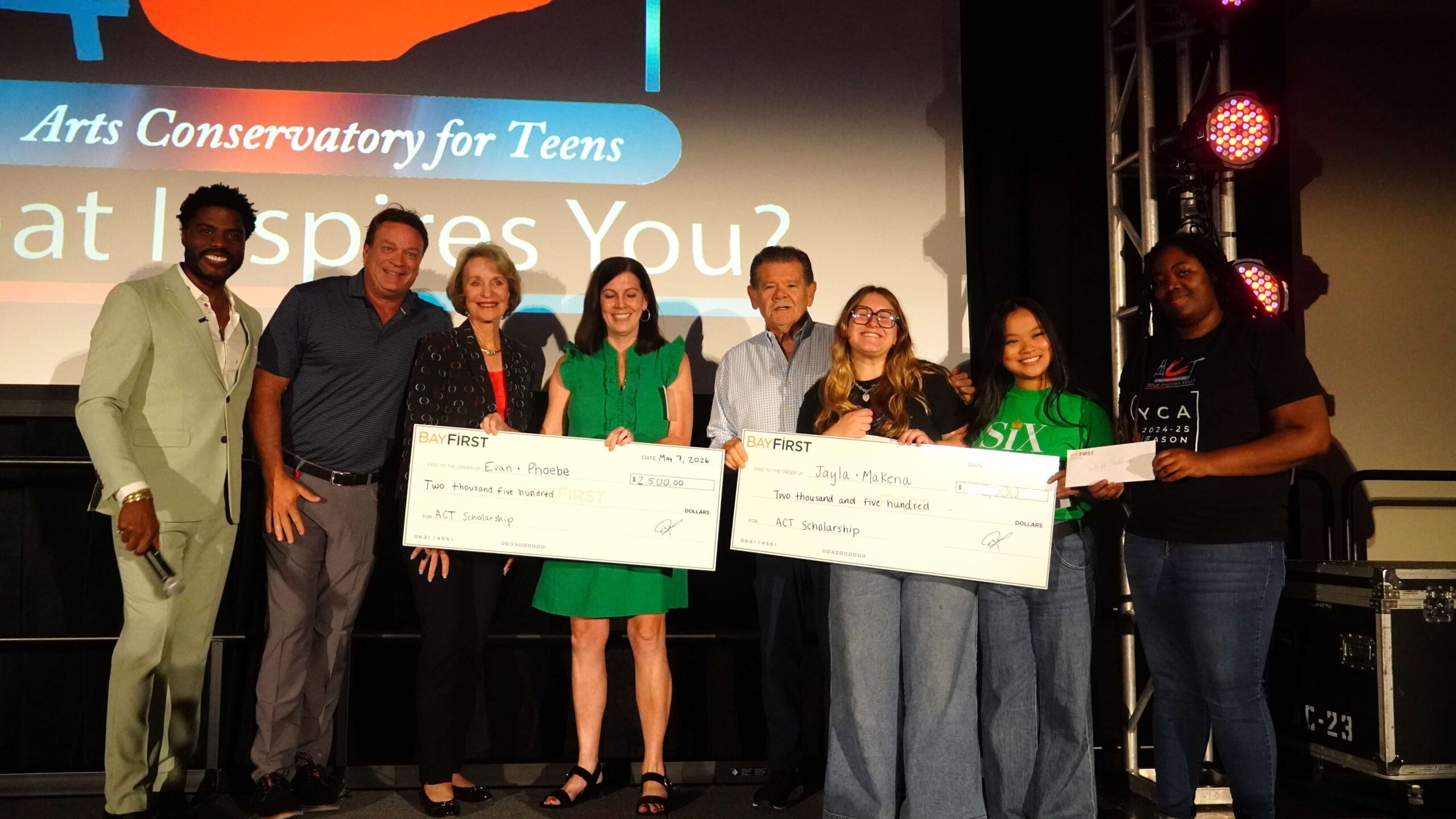 Eight people stand on a stage, some holding large scholarship checks, in front of a screen displaying "Arts Conservatory for Teens," celebrating achievements in art and painting.