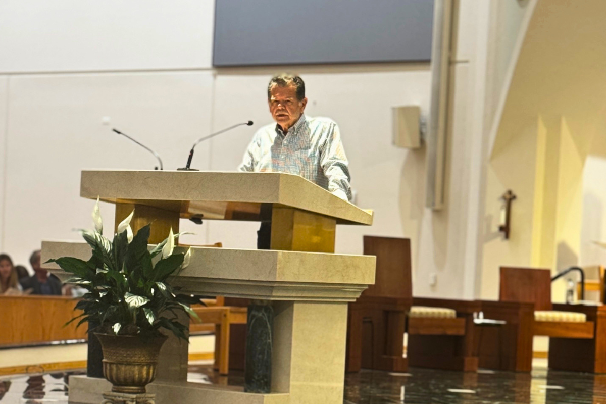 A man stands at a stone podium with microphones, speaking inside St. Petersburg Catholic High School’s church. A potted plant is placed in front of the podium as he announces new scholarships for students.