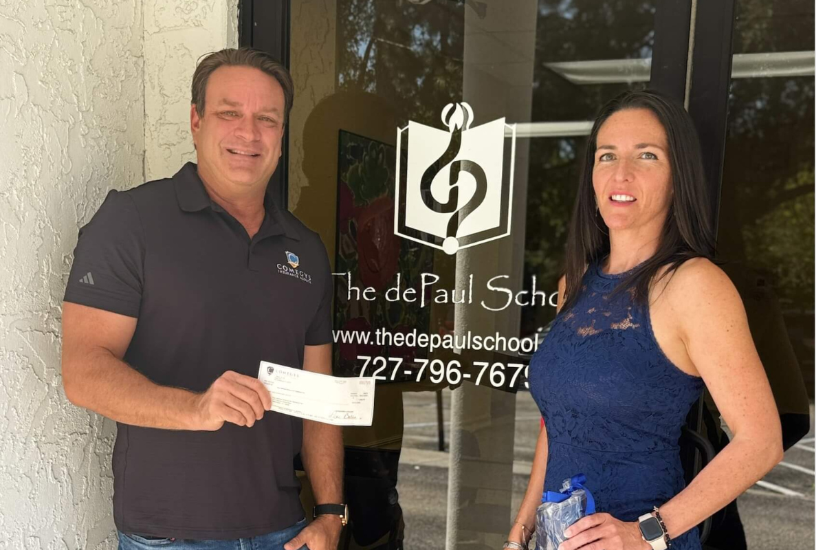 Two people stand in front of the dePaul School of Dyslexia entrance; the man on the left, representing Comegys Insurance Agency, holds a check as both face the camera.