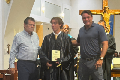 A young man in graduation robes holds a plaque, standing between two men inside St. Petersburg Catholic High School’s church with a large crucifix in the background, celebrating scholarships from Comegys Insurance Agency.
