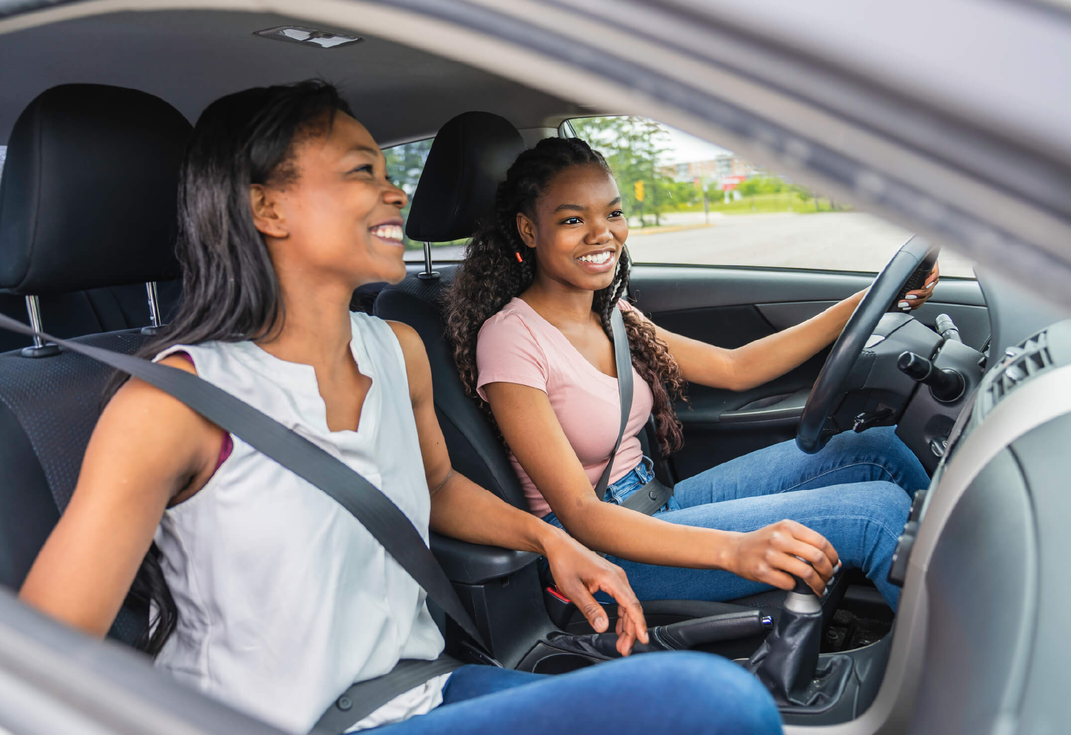 Two women sitting in a car; the driver smiles while holding the wheel and gear shift, and the passenger is also smiling. Both wear seatbelts, enjoying peace of mind unlike those with an underinsured home.