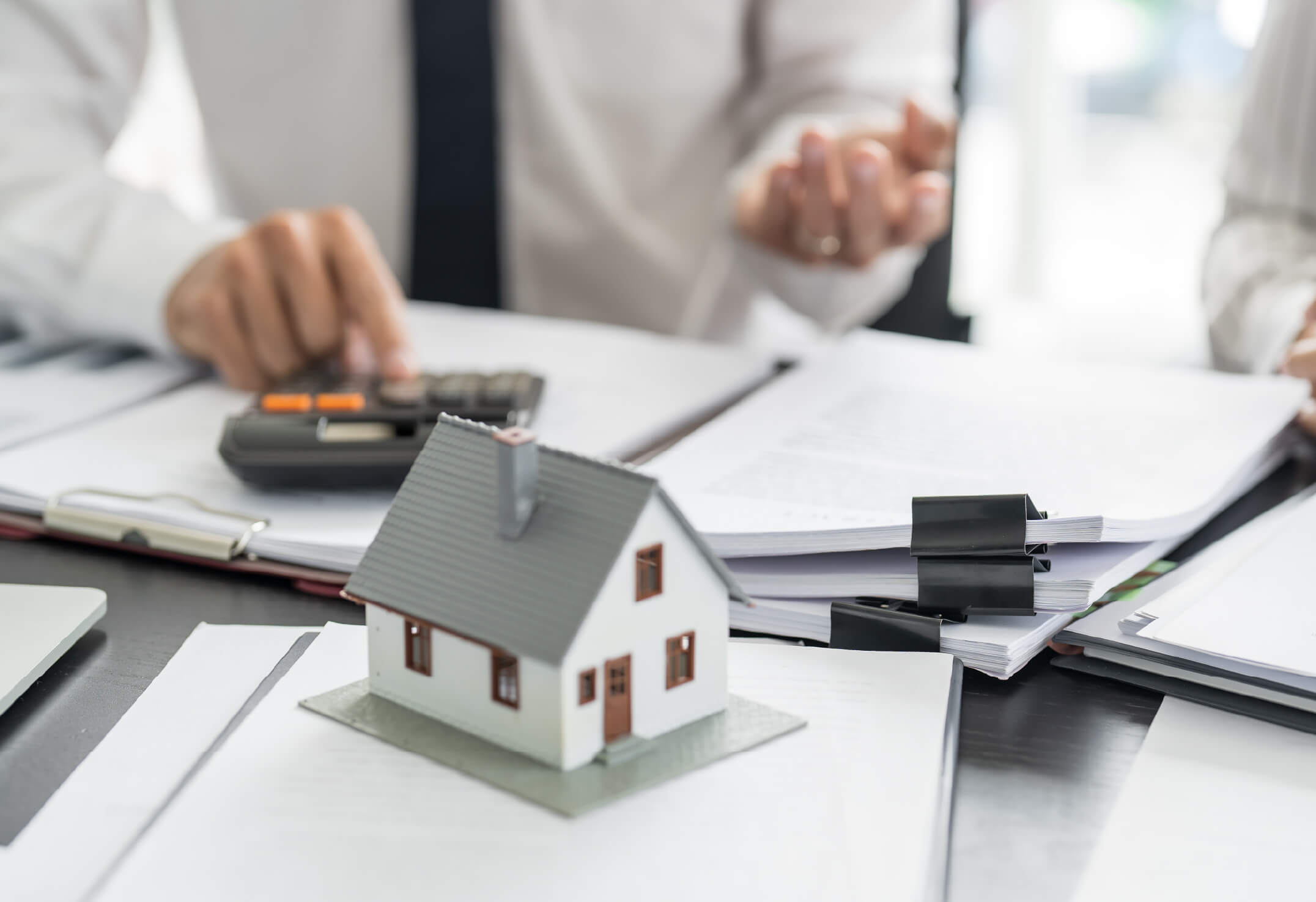A small model house sits on paperwork, while a person in business attire uses a calculator and gestures with their hand at a desk—highlighting concerns about an underinsured home.