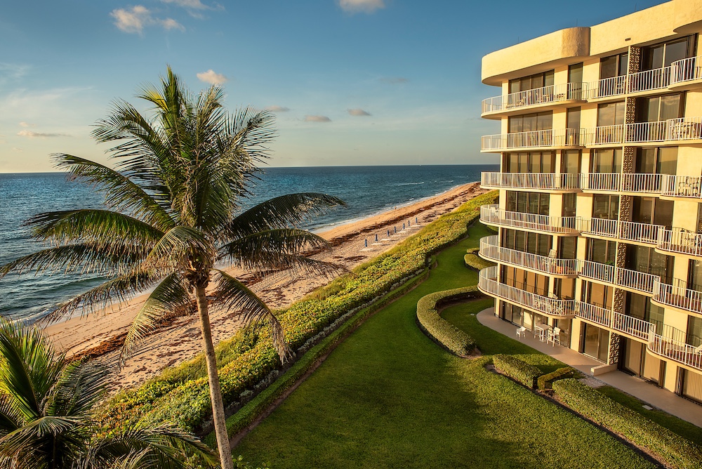 A modern beachfront building with balconies overlooks a grassy lawn, palm trees, and a sandy ocean shoreline at sunset—an ideal setting where homeowners insurance for condos ensures peace of mind for residents enjoying coastal living representing Homeowners Insurance for Condos in Florida